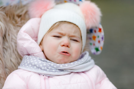 Sad crying hungry baby girl sitting in the pram or stroller on cold autumn, winter or spring day. Weeping child in warm clothes, fashion stylish baby coat and hat. Snow falling downの写真素材