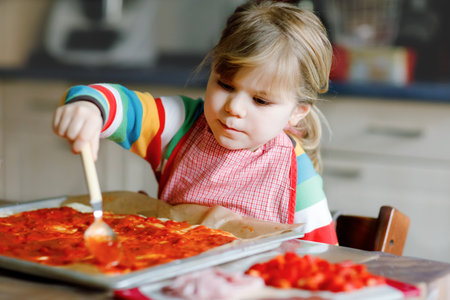 Adorable little toddler girl making italian pizza at home. Cute happy child having fun in home kitchen, indoors. Kid, preschooler helping and preparing healthy mealの写真素材