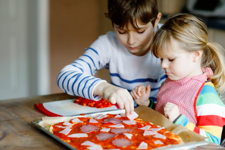 Two siblings, little children making italian pizza at home. Cute toddler girl and school boy having fun in home kitchen, indoors. Brother and sister, family helping and preparing mealの写真素材