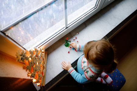 Litte toddler girl sitting by window and decorating small glass Christmas tree with tiny xmas toys. Happy healthy child celebrate family traditional holiday. Adorable baby.の写真素材