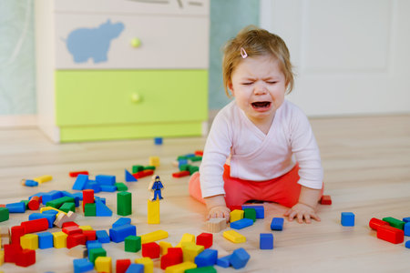 Upset crying baby girl with educational toys. Sad tired or hungry alone healthy child sitting near colorful different wooden blocks at home or nursery. Baby missing mother in daycareの写真素材