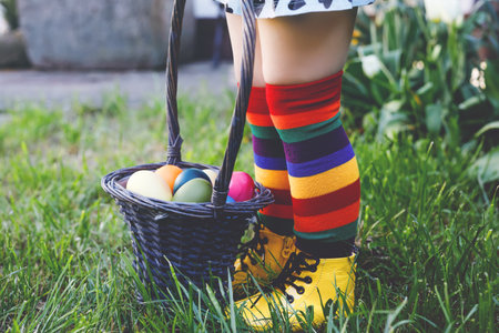 Close-up of legs of toddler girl with colorful stockings and shoes and basket with colored eggs. Child having fun with traditional Easter eggs hunt, outdoors. Celebration of christian holiday.の写真素材