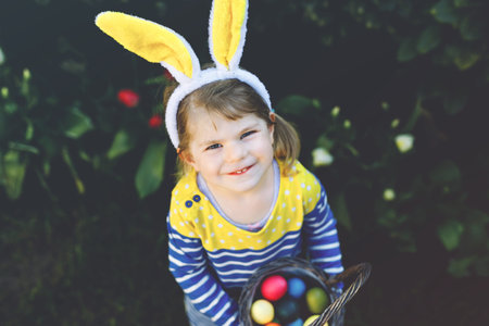 Cute little toddler girl with bunny ears having fun with traditional Easter eggs hunt on warm sunny...