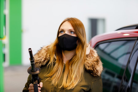 Woman wear medical mask at self-service gas station, hold fuel nozzle, refuel the car with petrol during corona virus pandemic lockdown. People in masks as preventive measure and covid protectionの写真素材