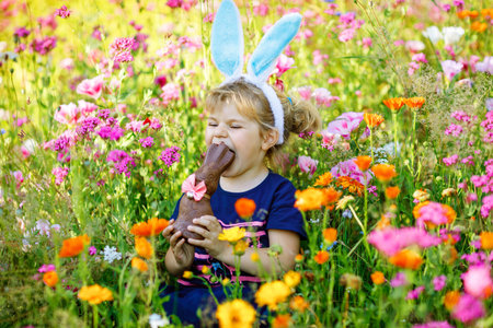 Portriat of adorable, charming toddler girl with Easter bunny ears eating chocolate bunny figure in...