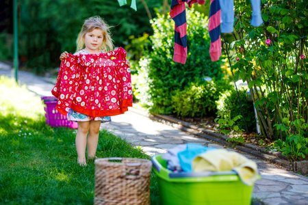 Toddler girl hanging clothes, fresh clean dresses and trousers for drying in garden, outdoors after making laundry. Happy child helping in household. Family working together, kid learning to help.の写真素材