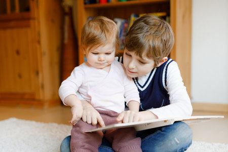 School kid boy reading book for little toddler baby girl, Two siblings sitting together and read books. Beautiful lovely family in love, cute baby and child having fun at home, indoors.の写真素材