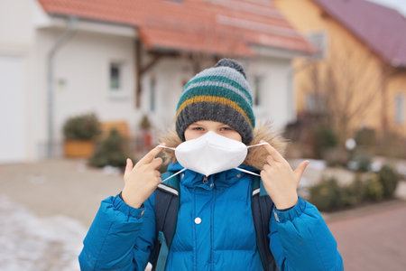 Kid boy wearing ffp medical mask on the way to school. Child backpack satchel. Schoolkid on cold autumn or winter day with warm clothes.の写真素材