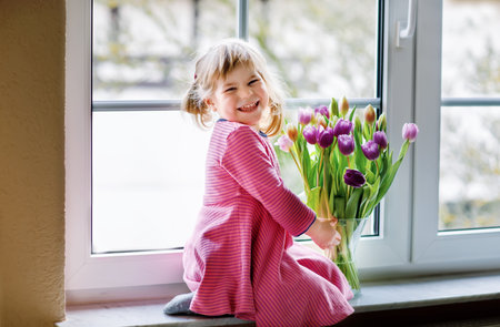 Little girl sitting by window with tulip flowers bouquet. Happy child, indoors. Mothers day, valentines day or birthday concept.の写真素材