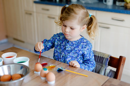 Excited little toddler girl coloring eggs for Easter. Cute happy child looking surprised at colorful colored eggs, celebrating holiday with family. Adorable kid at home with different bright colors.の写真素材
