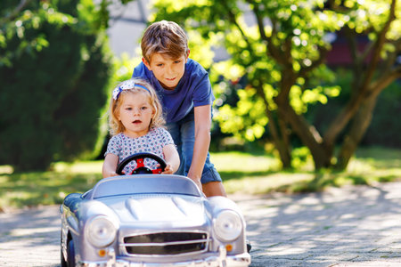 Two happy children playing with big old toy car in summer garden, outdoors. Boy driving car with little girl, cute sister inside. Laughing and smiling kids. Family, childhood, lifestyle concept.の写真素材