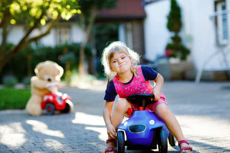 Little adorable toddler girl driving toy car and having fun with playing with plush toy bear, outdoors. Gorgeous happy healthy child enjoying warm summer day. Smiling stunning kid in gadenの写真素材