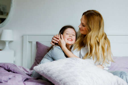 Lovely young mother and cute school kid girl cuddling together in bed in morning. Happy family of young single woman and beautiful daughter, indoors, getting ready and morning routine.の写真素材