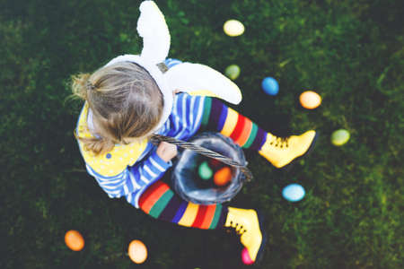 Close-up of legs of toddler girl with colorful stockings and shoes and basket with colored eggs. Child having fun with traditional Easter eggs hunt, outdoors. Unrecognizable face, no face.の写真素材