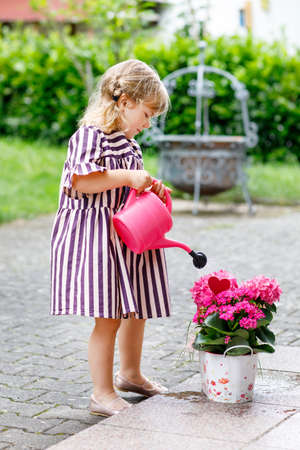 Portrait of adorable, charming toddler watering flowers with water can. Smiling happy child on summer day with colorful flowers, outdoors. Happiness and summertime. Blooming plants for mothers day.の写真素材