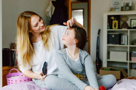 Loving mother brushing kid daughter hair sitting on bed, smiling single mom helping child girl with hairstyle at home getting ready, family care, morning preparations and routine lifestyle conceptの写真素材