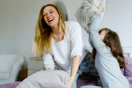Loving mother and school kid girl making pillow fight together. Happy family, woman and cute daughter having fun together in bed. Family care, morning routing and lifestyle.の写真素材