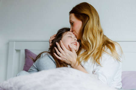Lovely young mother and cute school kid girl cuddling together in bed in morning. Happy family of young single woman and beautiful daughter, indoors, getting ready and morning routine.の写真素材