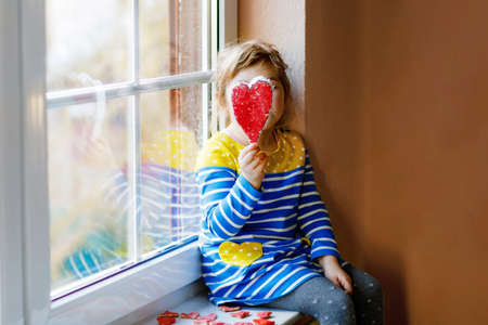 Cute little girl by window holding selfmade heart as gift for Valentines day, Mothers day or birthday. Adorable happy smiling child indoors.の写真素材