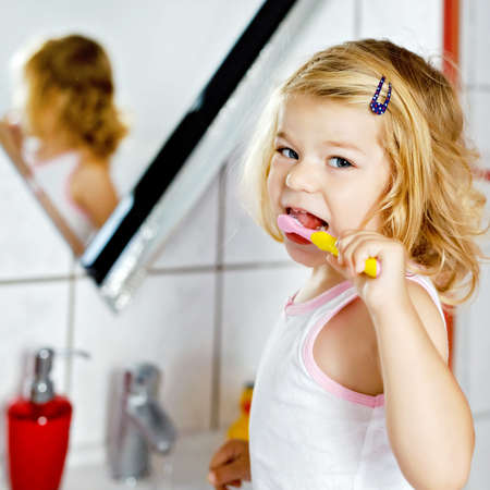 Cute adorable toddler girl holding toothbrush and brushing first teeth in bathroom after sleeping. Gorgeous baby child learning to clean milk tooth. Morning healthy hygiene routine for childrenの写真素材
