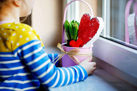 Little girl sitting by window with hyacinth flowers and hand-made heart. Happy child, indoors. Mothers day, valentines day or birthday and spring concept.の写真素材