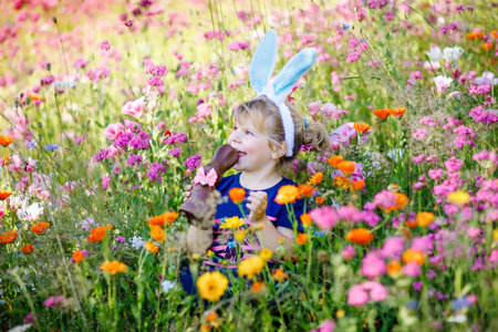 Portriat of adorable, charming toddler girl with Easter bunny ears eating chocolate bunny figure in...