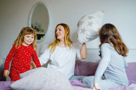 Loving mother and two little sisters girls making pillow fight together. Happy family, woman and cute daughters, toddler and school child having fun together in bed.の写真素材