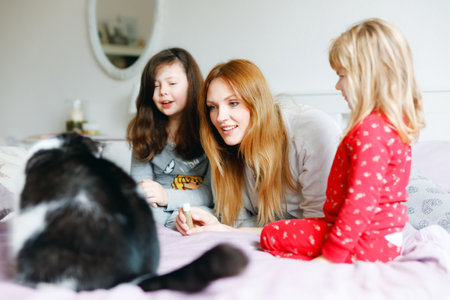 Lovely mother and two little girls daughters cuddling together with cat in bed in morning. Happy family of young single woman and sisters, toddler child and school girl, indoors.の写真素材