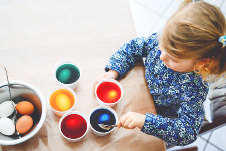 Closeup of little toddler girl coloring eggs for Easter. Close-up of child looking surprised at colorful colored eggs, celebrating holiday with family. From above, unrecognized face.の写真素材