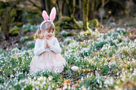 Little girl with Easter bunny ears making egg hunt in spring forest on sunny day, outdoors. Cute...