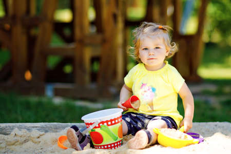 Cute toddler girl playing in sand on outdoor playground. Beautiful baby having fun on sunny warm summer sunny day. Happy healthy child with sand toys and in colorful fashion clothes.の写真素材