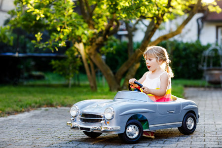 Little adorable toddler girl driving big vintage toy car and having fun with playing outdoors. Gorgeous happy healthy child enjoying warm summer day. Smiling stunning kid playing in domestic gardenの写真素材