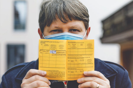 Man with medical mask holding German inernational certificate of the vaccination. He showing the information about his vaccine against covid 19 corona virus.の写真素材