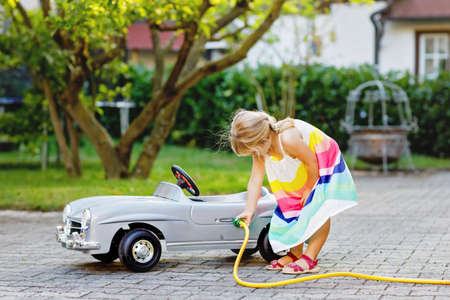 Little toddler girl playing with big vintage toy car and having fun outdoors in summer. Cute child refuel car with water. Girl using garden hose and fill up with gasoline, role game gas station.の写真素材