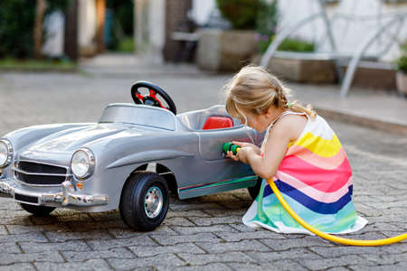 Little toddler girl playing with big vintage toy car and having fun outdoors in summer. Cute child refuel car with water. Girl using garden hose and fill up with gasoline, role game gas station.の写真素材
