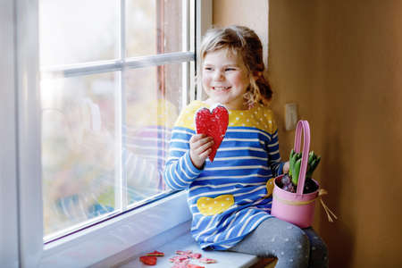 Little girl sitting by window with hyacinth flowers and hand-made heart. Happy child, indoors. Mothers day, valentines day or birthday and spring concept.の写真素材