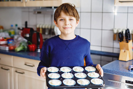 Happy blond school kid boy baking blueberry vanilla muffins in domestic kitchen, indoors. Funny lovely healthy child having fun with helping.の写真素材