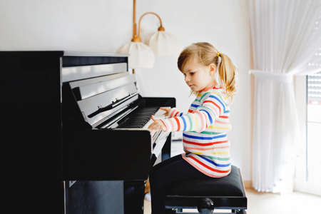Beautiful little toddler girl playing piano in living room. Cute preschool child having fun with learning to play music instrument with learning concept during homeschooling corona virus lockdown.の写真素材