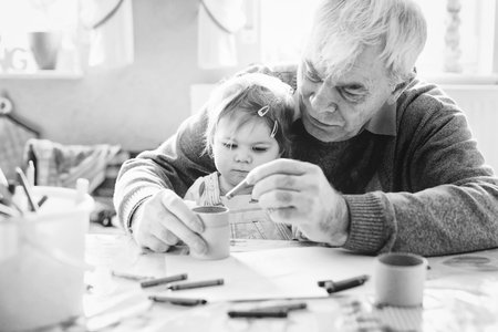 Cute little baby toddler girl and handsome senior grandfather painting with pencils at home. Grandchild and man having fun together. Family and generation in love. Old black and white imageの写真素材