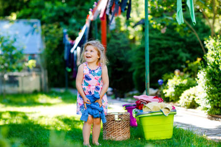 Toddler girl hanging clothes, fresh clean dresses and trousers for drying in garden, outdoors after making laundry. Happy child helping in household. Family working together, kid learning to help.の写真素材