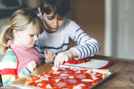 Two siblings, little children making italian pizza at home. Cute toddler girl and school boy having fun in home kitchen, indoors. Brother and sister, family helping and preparing mealの写真素材