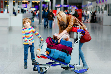 Two little kids, boy and girl, siblings and mother at the airport. Children, family traveling, going on vacation by plane and waiting on trolley with suitcases pushing by woman at terminal for flight.の写真素材