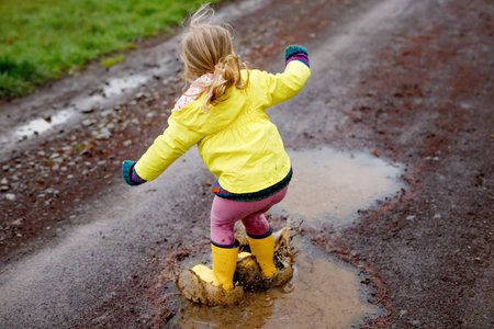 Little toddler girl wearing yellow rain gum boots, running and walking during sleet. Happy child in...