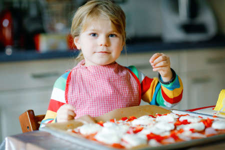 Adorable little toddler girl making italian pizza at home. Cute happy child having fun in home kitchen, indoors. Kid, preschooler helping and preparing healthy mealの写真素材
