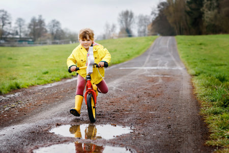 Little toddler girl wearing yellow rain gum boots, running with balance bike during sleet. Happy child driving, biking with bicycle into puddle, splashing water, outdoor activity. Happiness, childhoodの写真素材