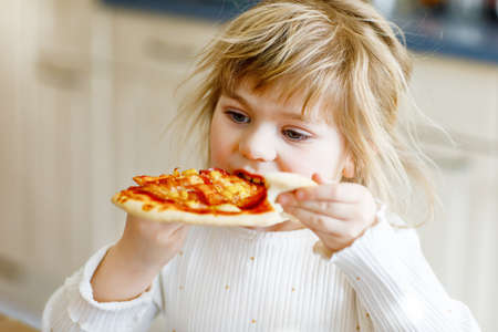 Adorable toddler girl eat italian pizza with vegetables and chees. Happy child eating fresh cooked healthy meal with tomatoes, corn and vegetables at home, indoors.の写真素材