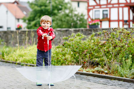 Little blond kid boy walking with big umbrella outdoors on rainy day. Portrait of cute preschool child having fun wear colorful waterproof clothes. Outdoor leisure walk on bad weather day with kidsの写真素材