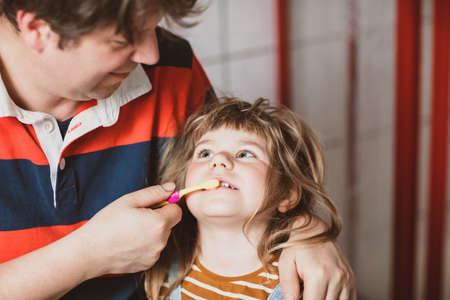 Father helping his little daughter with brushing teeth. Little toddler girl and dad in bathroom, making hygiene actions. Happy family, man and child. Morning routine with children.の写真素材