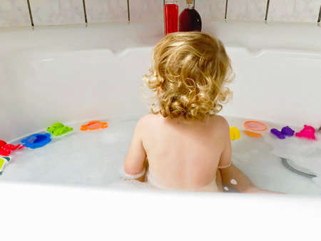 Little toddler girl taking bath in bathtub. Healthy baby child playing with rubber gum toys and having fun. Washing, cleaning, hygiene for children. From back, unrecognizable personの写真素材