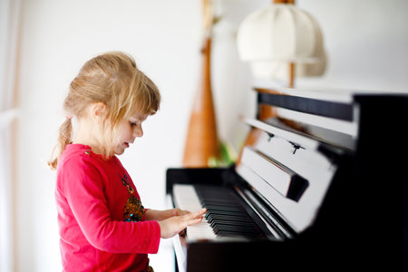 Beautiful little toddler girl playing piano in living room. Cute preschool child having fun with learning to play music instrument. Early musical education for children.の写真素材